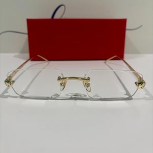 Puede incluir: Gafas sin montura con monturas doradas y detalles verdes en las patillas. Las gafas tienen una caja roja rectangular detrás y el fondo tiene el texto "WHERE LIFE BEGINS AND".