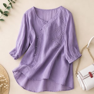 May include: A light purple blouse featuring a diagonal button placket, three-quarter length sleeves with buttoned cuffs, and a delicate floral embroidery pattern on the right side. The garment has a round neckline and a curved hemline.