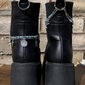 May include: Pair of black platform boots with decorative silver chains and charms. The boots have a chunky sole and a ribbed knit upper. The chains are adorned with colorful beads and small charms. The boots are set against a brick wall.