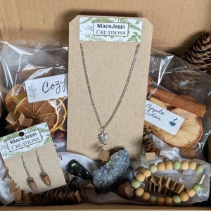 May include: An open box filled with various items, including a silver necklace with a leaf pendant, earrings, a bracelet, dried orange slices, cinnamon sticks, and a pine cone. The box contains the text "MacnJessi Creations" on the tags.