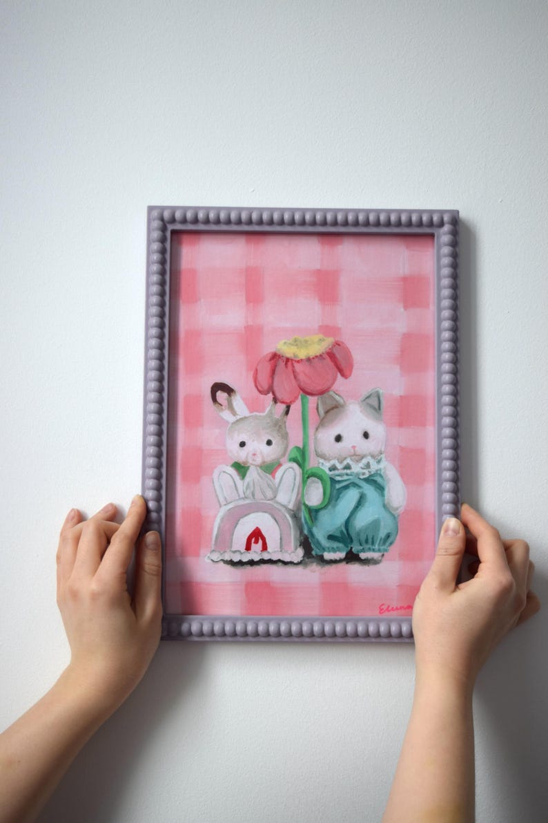 Peut inclure: Tableau encadr&eacute; repr&eacute;sentant un lapin et un chat, sur fond rose &agrave; carreaux. Le lapin porte du rose, le chat du bleu. Le cadre est violet clair. L'&oelig;uvre a un style fantaisiste et illustratif.