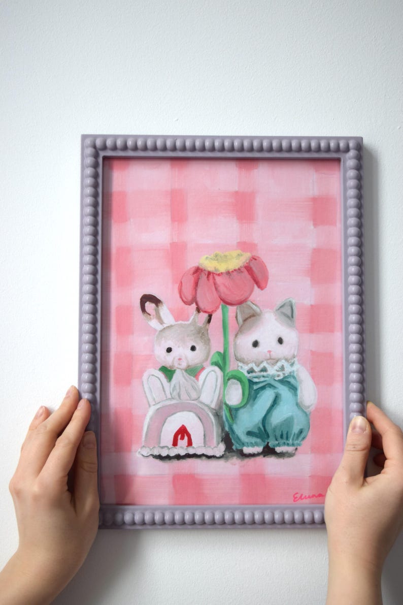 Peut inclure: Peinture encadr&eacute;e repr&eacute;sentant deux personnages de dessins anim&eacute;s, un lapin et un chat, pr&egrave;s d'une grande fleur rose. Le fond est un motif &agrave; carreaux roses et blancs. Le cadre est de couleur violet clair.