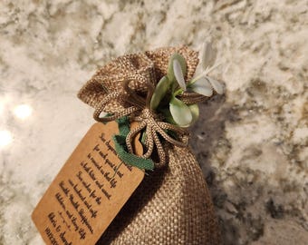 May include: A small burlap bag tied with twine and adorned with a sprig of greenery and a white flower. A brown tag is attached, bearing handwritten text. The tag reads "Five sugared almonds for each guest to eat..." and is personalized with a date.