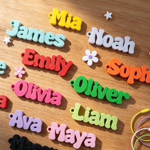 Puede incluir: Una colección de etiquetas de nombre de plástico tridimensionales y coloridas con los nombres Mia, James, Noah, Emily, Sophia, Olivia, Oliver, Liam, Ava, Maya y Mason, acompañadas de pequeñas formas de estrellas y flores y varios anillos metálicos.