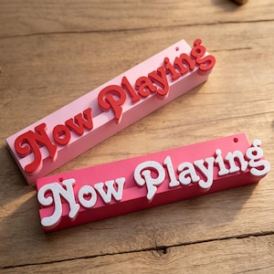 May include: Two rectangular display stands featuring the text Now Playing in raised, stylized lettering. The top stand is light pink with red text, while the bottom stand is dark pink with white, glittery text. Both have a small hole on the right.