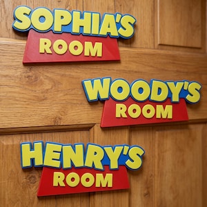 Puede incluir: Tres letreros de puerta personalizados con letras amarillas y bordes azules sobre una base trapezoidal roja. Los letreros muestran el texto SOPHIA'S ROOM, WOODY'S ROOM y HENRY'S ROOM en una fuente gruesa, montados en una puerta de madera.