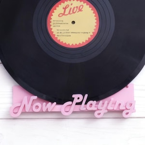 Puede incluir: Un disco de vinilo negro con una etiqueta amarilla y roja "Live" se muestra en un soporte rosa "Now Playing". El disco está sobre una superficie de madera blanca.
