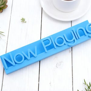 Puede incluir: Un letrero tridimensional de color azul brillante con el texto "Now Playing" en una fuente sans-serif. Las letras están realzadas sobre una base rectangular, creando una pieza de exhibición moderna y llamativa con un acabado mate.