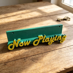 May include: A green horizontal display stand featuring the yellow raised text "Now Playing" in a cursive font. The stand has a flat ledge for holding items and two circular mounting holes along the top back edge for wall installation.