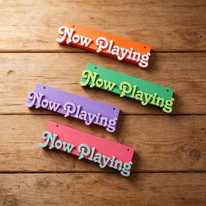 May include: Four brightly coloured wooden signs, each reading "Now Playing" in a decorative font. The signs are orange, green, purple, and pink, with contrasting text. They are displayed on a wooden surface.