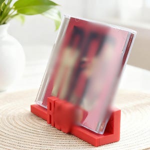May include: A red CD holder displaying a CD in a clear plastic case. The holder is made of solid red material and angles the CD for display. A white vase with a green plant is in the background.