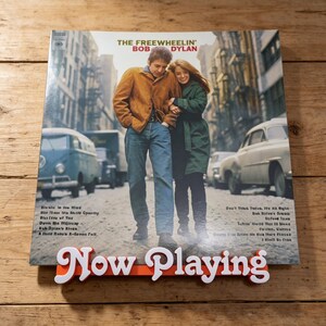 May include: A vinyl record cover for The Freewheelin' Bob Dylan featuring a couple walking down a city street, displayed on an orange and white Now Playing stand. The cover shows a man in a tan jacket and blue jeans with a woman in a green coat.