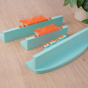 May include: Three teal-colored wooden display shelves with orange and teal lettering. The shelves are arranged on a light wood surface. A white pot with green leaves is in the background.