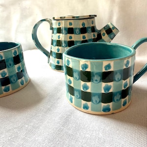 May include: A ceramic tea set consisting of a teapot and two mugs, each featuring a hand-painted checkered pattern in shades of teal, light blue, and dark green, with small blue floral motifs centered within each light blue square of the grid.