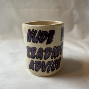 May include: A beige ceramic mug with the words "NUDE READING ADVICE" in purple lettering. The mug has a cylindrical shape and a slightly rough texture, suggesting a handmade quality.