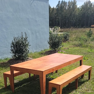 May include: A wooden table and two benches set on a grassy lawn. The table and benches are made of light brown wood with a natural grain pattern. The table is rectangular and has four legs. The benches are also rectangular and have four legs. The table and benches are in front of a blue wall.