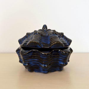 May include: A ceramic trinket box with a lid, featuring a seashell design. The box is glazed in a glossy, dark blue with brown accents, and sits on a light-colored surface. The box has a textured, wavy pattern.