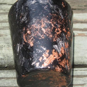 Può includere: Un bicchiere nero, cilindrico, con dettagli color rame. Il bicchiere ha una superficie strutturata e una parte superiore leggermente svasata. I dettagli in rame hanno forme irregolari e aggiungono interesse visivo al pezzo.