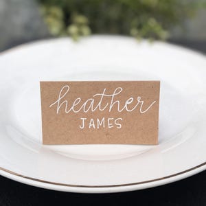May include: A brown card with white calligraphy that reads "heather JAMES". The card is sitting on a white plate.