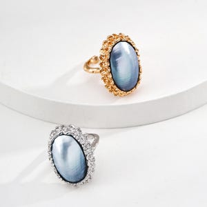 Peut inclure: Deux bagues avec des pierres ovales bleu-gris iridescentes. L'une a une monture dorée et un sertissage doré texturé. L'autre a une monture argentée et un sertissage argenté avec de petites pierres claires.
