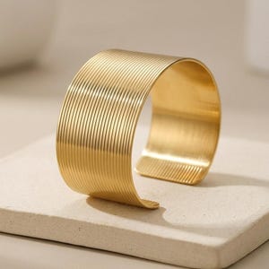 Peut inclure: Un bracelet manchette doré avec un motif strié. Le bracelet ouvert a une forme incurvée et repose sur une surface de couleur claire. La surface du bracelet présente une série de lignes parallèles.