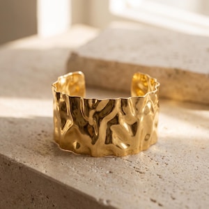 Puede incluir: Un brazalete dorado con un diseño texturizado y orgánico. El brazalete abierto tiene un borde ondulado y una apariencia martillada. La joya se exhibe sobre una superficie de tonos neutros.