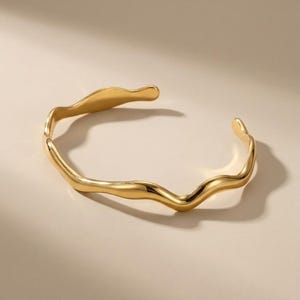 Può includere: Un bracciale a polsino color oro con un design fluido e organico. Il bracciale aperto ha una superficie ondulata e strutturata, creando un'estetica scultorea e fluida. Questo gioiello è un accessorio di tendenza.