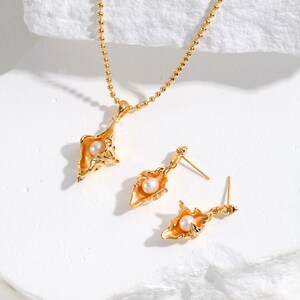 Könnte beinhalten: Ein goldfarbenes Schmuckset mit einer Halskette und einem Paar Ohrringen. Die Halskette hat eine zarte Kette mit einem Anhänger, und die Ohrringe sind als Stecker ausgeführt. Jedes Stück hat ein goldfarbenes, durchbrochenes Design mit einer Perle in der Mitte.