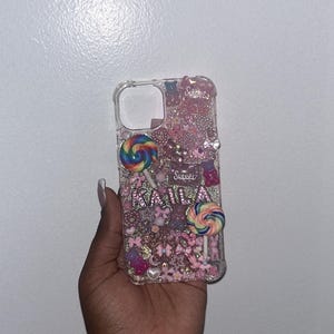 Può includere: Una custodia per telefono trasparente con uno sfondo rosa e scintillante. La custodia è decorata con caramelle colorate, glitter e il nome "Kaila" in lettere rosa. Il testo "Sweet" è incluso anche in lettere rosa.