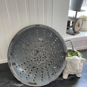 Op de afbeelding: Een vintage grijze emaille zeef met een gespikkelde afwerking en roestvlekken. De zeef heeft een ronde vorm en een handvat aan de zijkant. Hij staat op een grijs aanrecht.
