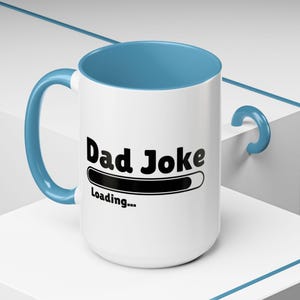 May include: White ceramic mug with a blue interior and handle. The mug features the text "Dad Joke Loading..." in black. The mug is on a white surface.