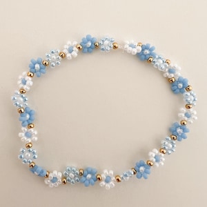Könnte beinhalten: Ein zartes blaues und weißes Perlenarmband mit kleinen blütenförmigen Perlen. Das Armband ist mit winzigen goldenen Perlen zwischen den Blütenperlen gefertigt.