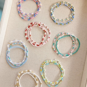 May include: A collection of beaded bracelets in various colors, including pink, blue, white, and green. Each bracelet features a daisy flower design. The bracelets are arranged on a light beige surface.