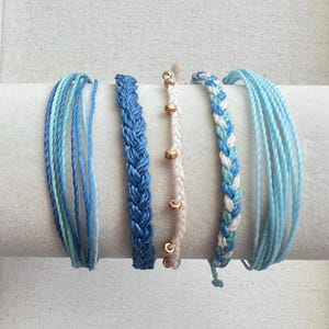 May include: A set of five bracelets featuring various designs: two multi-strand bracelets in shades of blue, one thick braided blue bracelet, one white cord bracelet with small gold-toned beads, and one braided bracelet in blue and white.