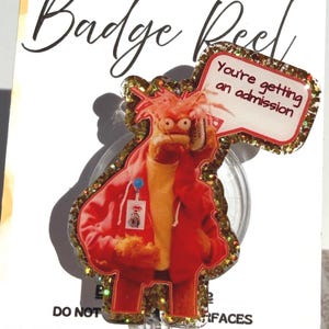 May include: A badge reel featuring a cartoon character in a red jacket holding a sign that says "You're getting an admission." The reel has a gold glitter border and is attached to a white card with text.