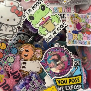 May include: A collection of colorful acrylic charms featuring various graphic designs including a green frog, a cartoon cat, a group of four women, Spongebob SquarePants, and text-based slogans like Jesus fills my cup and You pose we expose.