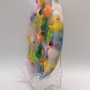 K&ouml;nnte beinhalten: Ein klares, ovales Kunstwerk aus Harz, gef&uuml;llt mit bunten Miniatur-Tierfiguren, darunter Enten, Fr&ouml;sche und andere Kreaturen. Das St&uuml;ck wird auf einem transparenten Kunststoffst&auml;nder pr&auml;sentiert.
