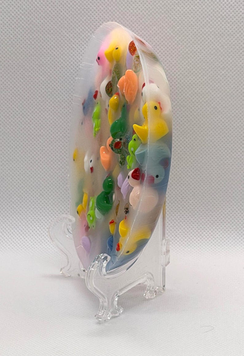 K&ouml;nnte beinhalten: Ein klares, ovales Kunstwerk aus Harz, gef&uuml;llt mit bunten Miniatur-Tierfiguren, darunter Enten, Fr&ouml;sche und andere Kreaturen. Das St&uuml;ck wird auf einem transparenten Kunststoffst&auml;nder pr&auml;sentiert.