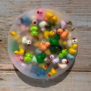 May include: A clear, round soap bar filled with colorful miniature ducks and frogs. The soap contains yellow, orange, green, white, and pink figurines. The soap is on a wooden surface.