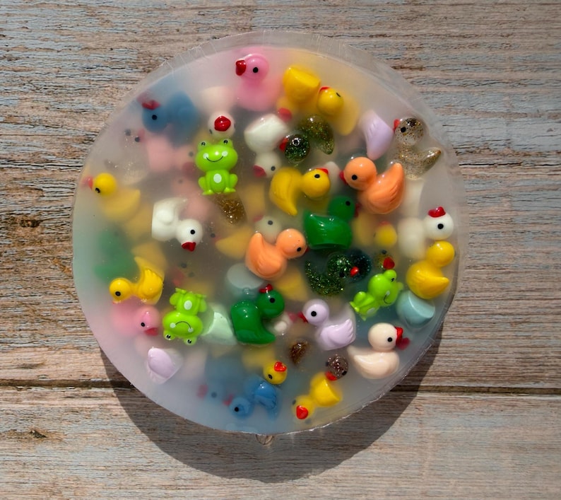 K&ouml;nnte beinhalten: Eine runde, transparente Seife gef&uuml;llt mit bunten Miniatur-Enten und Fr&ouml;schen. Die Seife enth&auml;lt gelbe, orangefarbene, gr&uuml;ne, wei&szlig;e und rosa Figuren. Die Seife liegt auf einer Holzoberfl&auml;che.