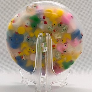 K&ouml;nnte beinhalten: Ein klares, rundes Kunstwerk aus Kunstharz, gef&uuml;llt mit zahlreichen Miniatur-Gummienten in verschiedenen Farben, darunter Gelb, Blau, Rosa und Gr&uuml;n. Die Enten sind in transparentem Kunstharz eingeschlossen und bilden ein einzigartiges Dekorationsst&uuml;ck. Pr&auml;sentiert auf einem transparenten Kunststoffst&auml;nder.
