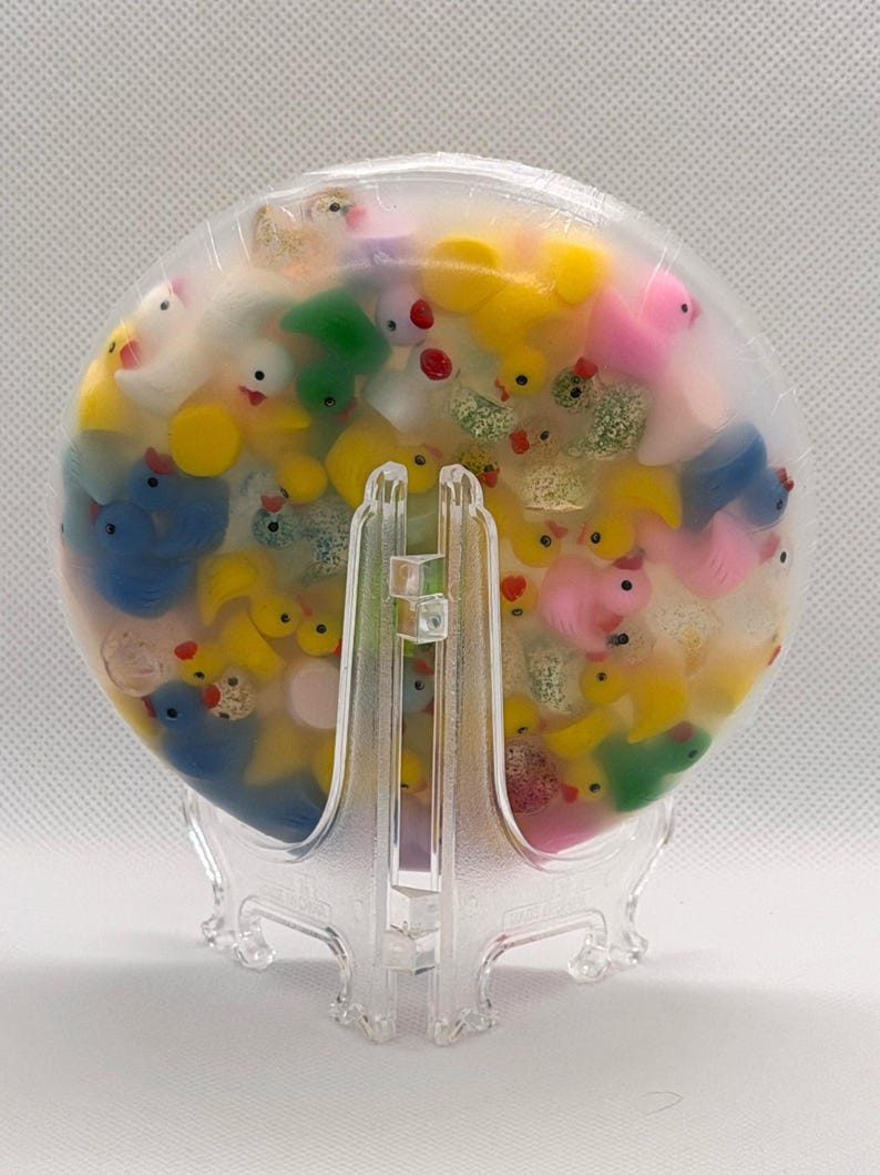 K&ouml;nnte beinhalten: Ein klares, rundes Kunstwerk aus Kunstharz, gef&uuml;llt mit zahlreichen Miniatur-Gummienten in verschiedenen Farben, darunter Gelb, Blau, Rosa und Gr&uuml;n. Die Enten sind in transparentem Kunstharz eingeschlossen und bilden ein einzigartiges Dekorationsst&uuml;ck. Pr&auml;sentiert auf einem transparenten Kunststoffst&auml;nder.