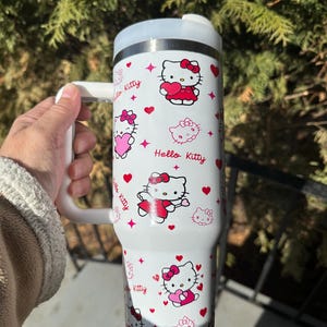 May include: White insulated tumbler featuring a repeating pattern of Hello Kitty characters, hearts, and stars. The tumbler has a white handle and a silver rim. The words "Hello Kitty" are printed on the tumbler.