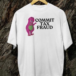 Könnte beinhalten: Weißes T-Shirt mit einem lila Dinosaurier-Charakter und dem Text "COMMIT TAX FRAUD" auf der Vorderseite.