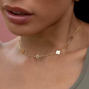 May include: A delicate gold necklace featuring a series of small charms. The charms include a square, a four-leaf clover, and a flower. The necklace is worn on a person's neck, and the background is blurred.