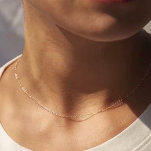 Puede incluir: Un delicado collar de oro sobre el cuello de una persona. El collar es una fina cadena con pequeños eslabones espaciados. La persona lleva una camiseta blanca. La iluminación es suave, resaltando el brillo sutil del collar.