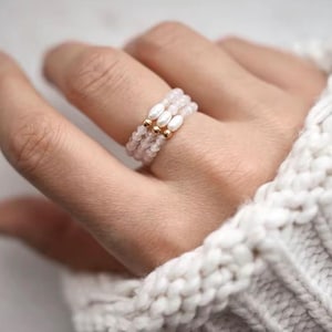 Puede incluir: Un delicado anillo con tres hilos de pequeñas cuentas de color rosa claro. Cada hilo está adornado con una cuenta dorada y una perla ovalada blanca. El anillo se lleva en un dedo, con un suave jersey blanco tejido en el fondo.