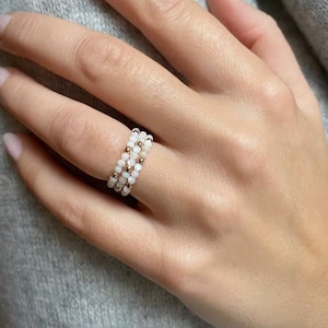 Puede incluir: Un conjunto de anillos apilables con tres anillos. Cada anillo está adornado con pequeñas cuentas blancas e intercalado con cuentas de color dorado. Los anillos se muestran en un dedo sobre un fondo gris suave.