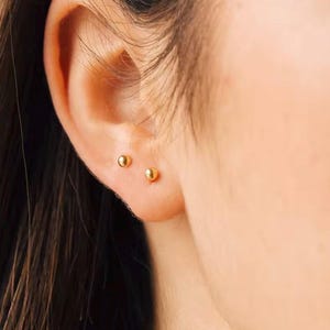 Puede incluir: Primer plano de la oreja de una persona con dos pequeños pendientes de bola de oro.