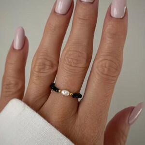 Puede incluir: Un anillo de cuentas negras con una perla blanca central y detalles dorados. El anillo se lleva en un dedo con esmalte de uñas rosa pálido. Es una pieza de joyería.
